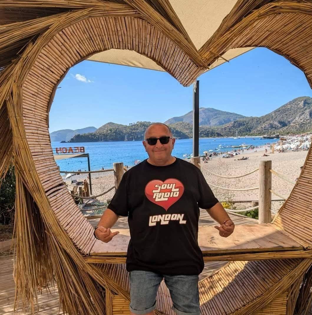 Man in sunglasses posing inside a heart-shaped bamboo frame with a beach and mountains in the background.