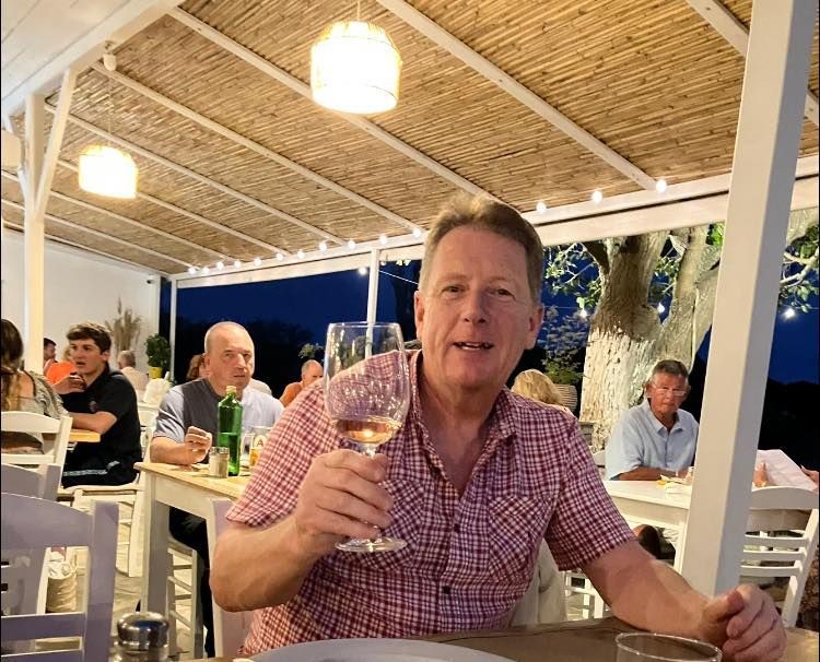 Man holding a glass of wine in an outdoor restaurant at night with other diners in the background.