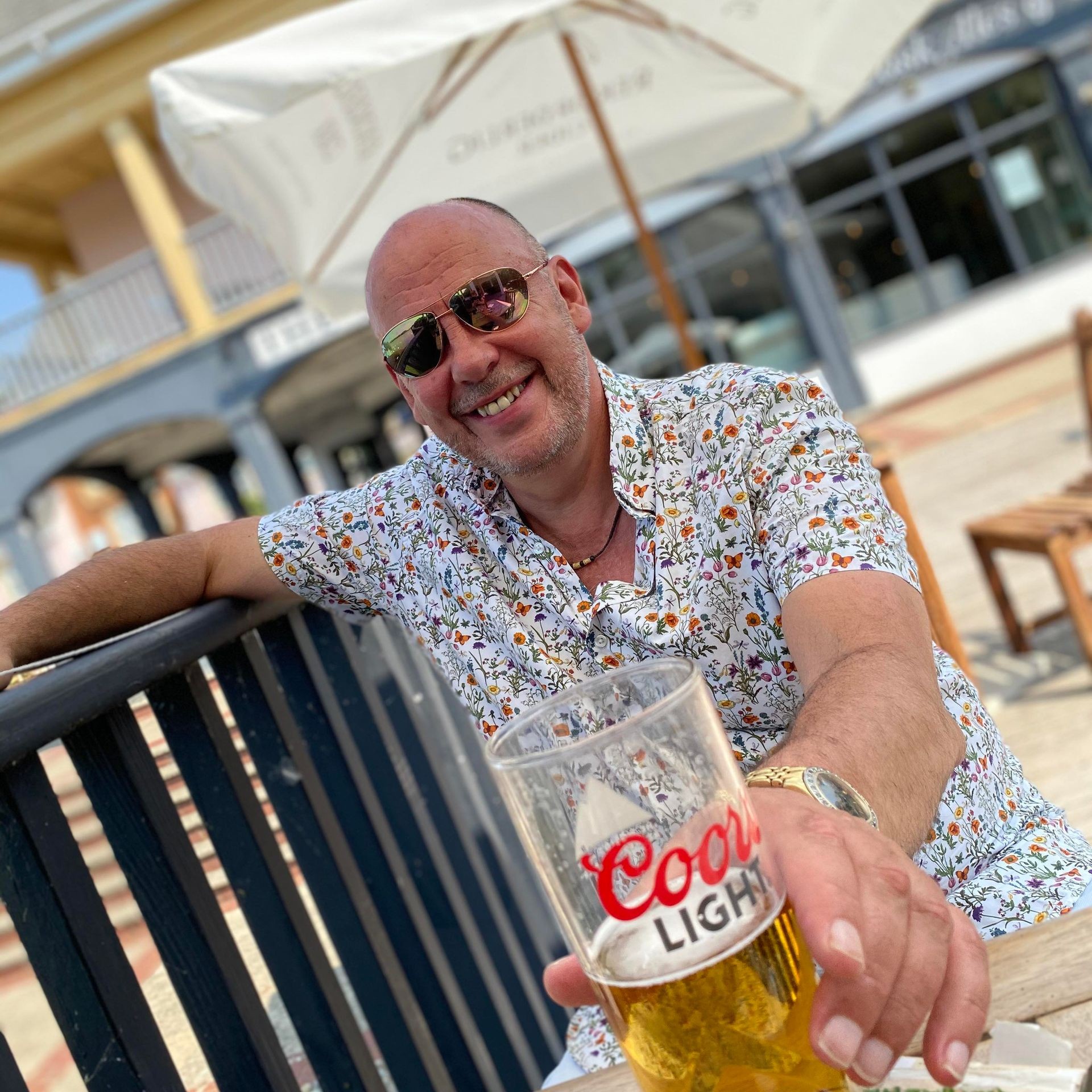 Person wearing a floral shirt holding a glass of Coors Light beer while sitting outdoors.