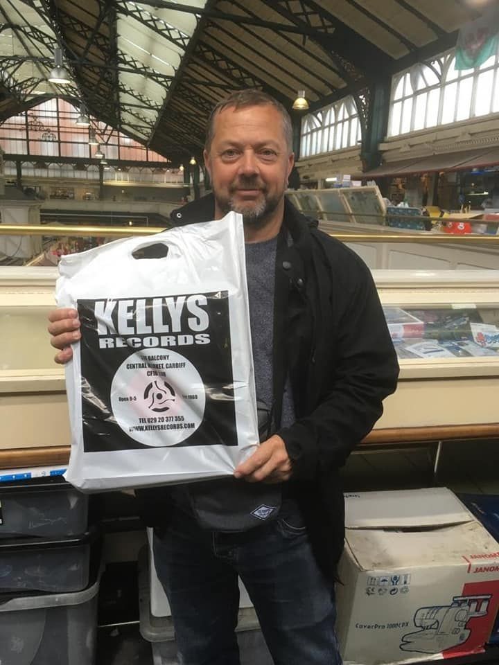 Person holding a white Kelly's Records shopping bag inside a market hall with an arched ceiling.