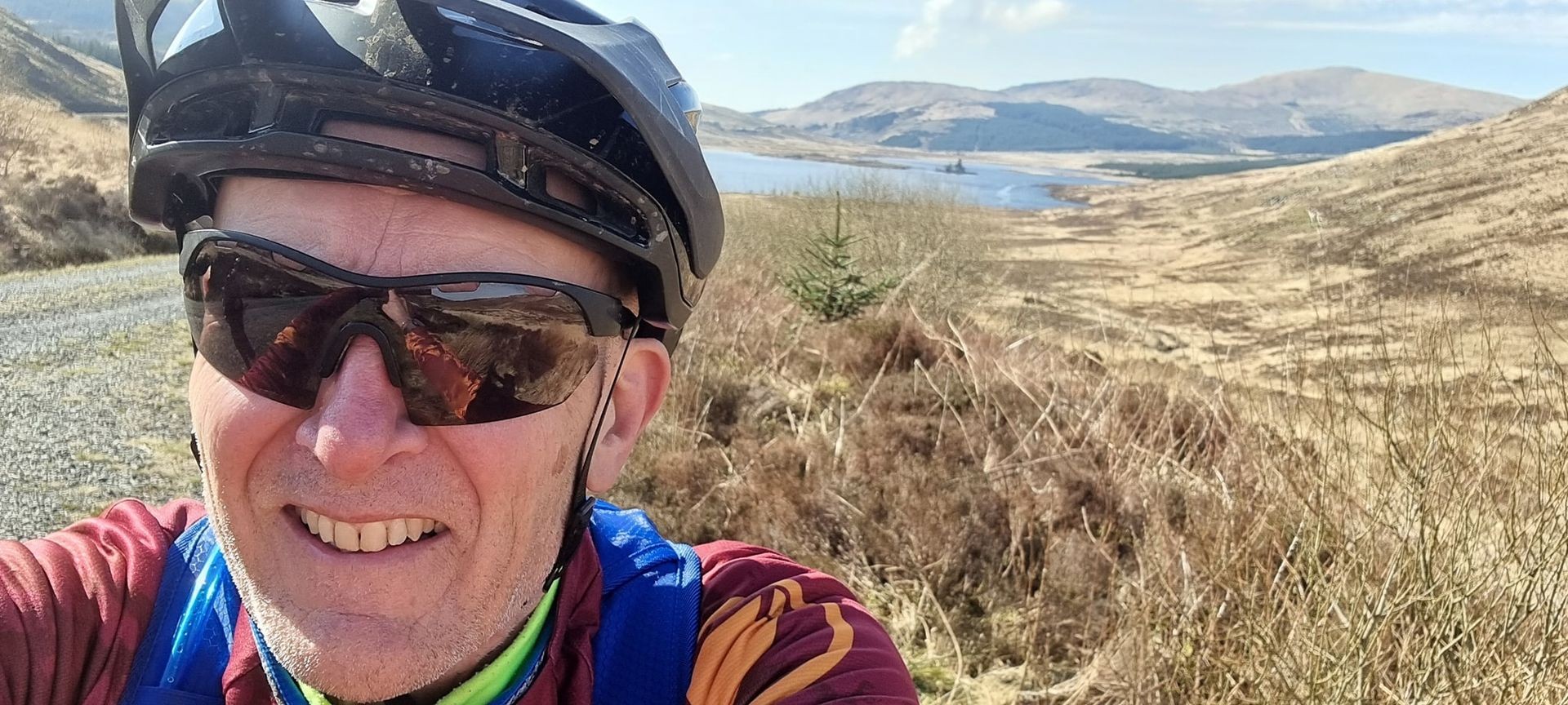 Person wearing a bicycle helmet standing on a trail with a scenic background of hills and a lake.