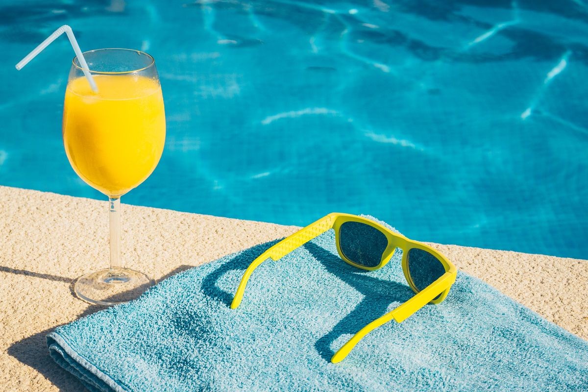 Yellow sunglasses on a blue towel and a glass of orange fresh water near the pool, summer holidays